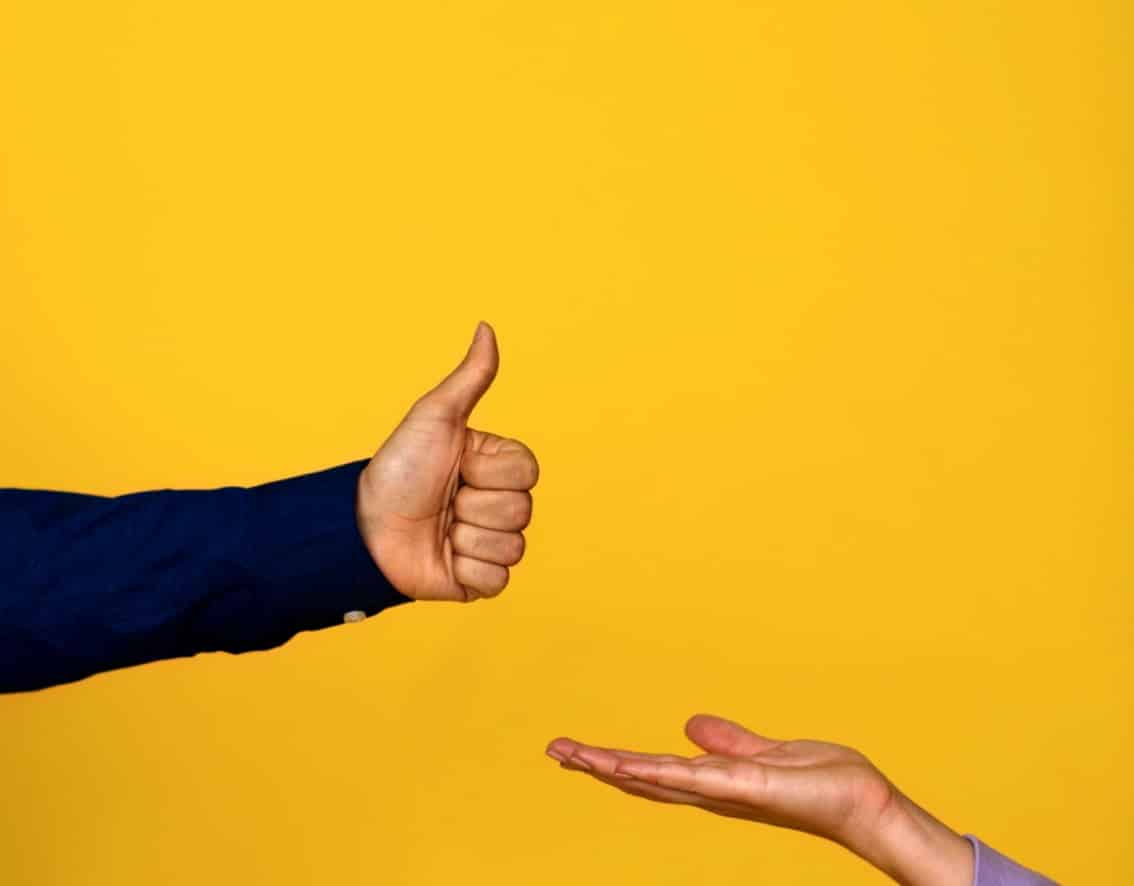 A person giving a thumbs-up gesture with one hand, while another persons open hand is extended below, both against a plain yellow background.